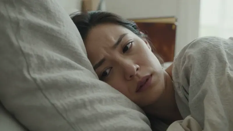 Mujer recostada sobre una almohada de lino arrugada en un dormitorio, con una expresión pensativa y la mirada enfocada, posiblemente reflexionando sobre sueños olvidados. Un diario de cuero desgastado se vislumbra en el fondo.