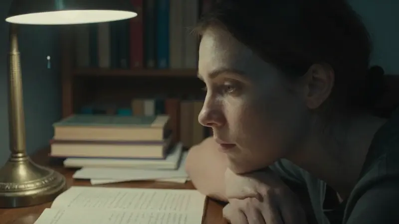 Mujer pensativa en un estudio con estanterías llenas de libros, iluminada por una lámpara antigua, rodeada de páginas de un diario de sueños.