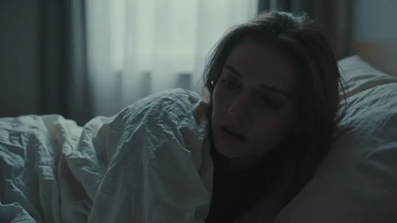 Mujer con expresión ansiosa en un dormitorio tenuemente iluminado, con sábanas revueltas y la luz de la mañana filtrándose a través de las cortinas.