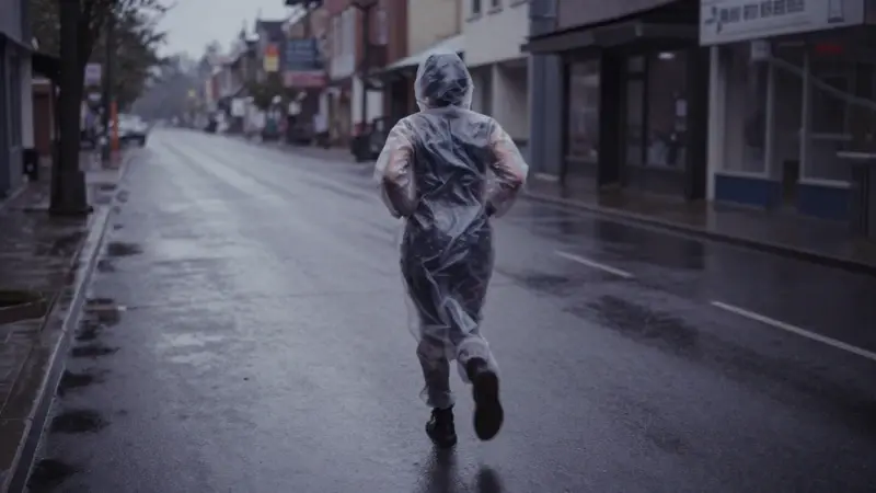 Figura solitaria corriendo por una calle desierta y mojada bajo la lluvia, con la ropa translúcida adherida al cuerpo y una atmósfera melancólica y amenazante.