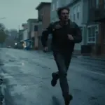 Figura solitaria corriendo por una calle desierta y lluviosa, con edificios silueteados al fondo y una expresión de intenso miedo.