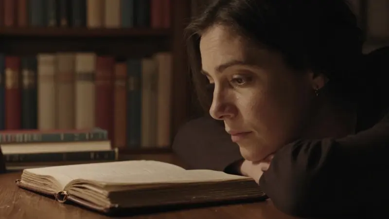 Mujer pensativa en un estudio con estanterías llenas de libros, iluminada tenuemente y con un diario abierto sobre un escritorio de madera.