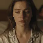 Mujer pensativa en la cama por la noche, con una figura borrosa en el fondo, evocando la reflexión sobre relaciones y sueños.