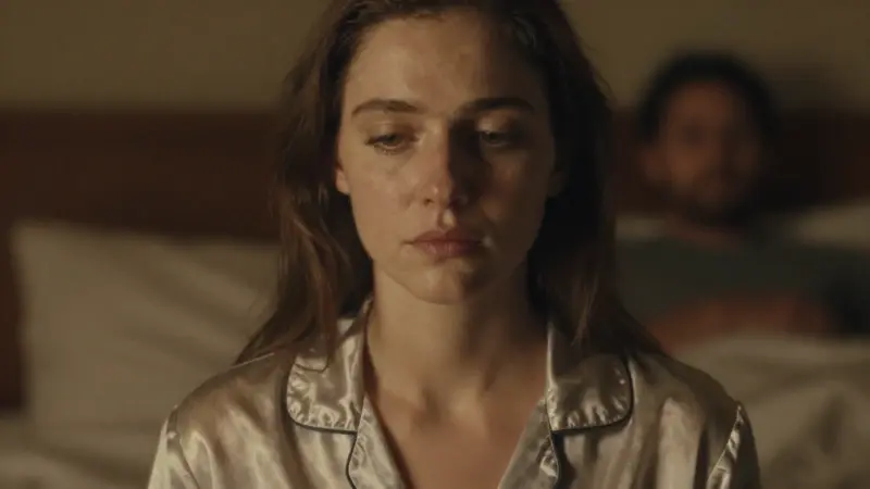 Mujer pensativa en la cama por la noche, con una figura borrosa en el fondo, evocando la reflexión sobre relaciones y sueños.