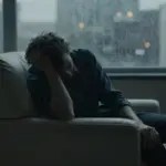 Persona sentada en un sillón en un consultorio tenuemente iluminado, con lluvia en la ventana y luces borrosas de la ciudad al fondo.