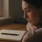 Mujer pensativa en una habitación tenuemente iluminada, con un diario de sueños y bolígrafo sobre una mesa de madera.