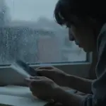 Mujer mirando una fotografía borrosa a través de una ventana lluviosa, en un dormitorio tenuemente iluminado con diarios de sueños dispersos, transmitiendo una atmósfera melancólica y pensativa.