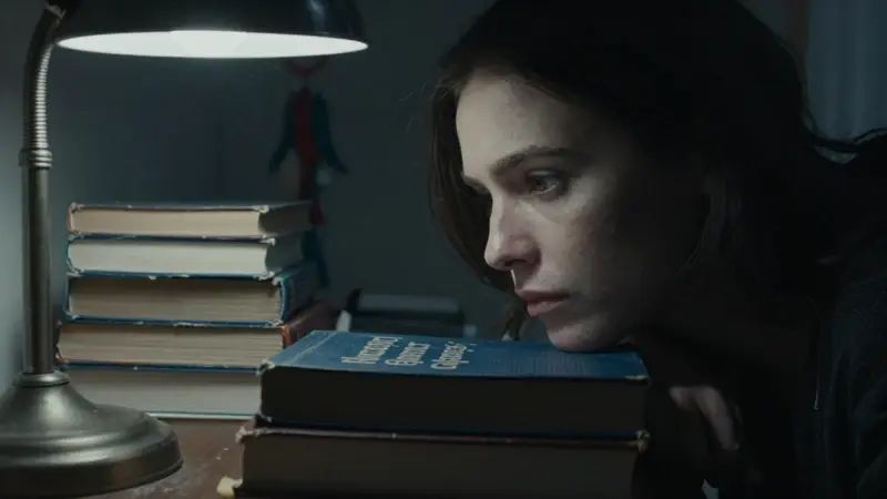 Primer plano de una mujer concentrada en un estudio tenuemente iluminado, rodeada de libros de psicología y una lámpara antigua sobre un escritorio. El ambiente es melancólico e introspectivo, con un atrapasueños borroso al fondo.