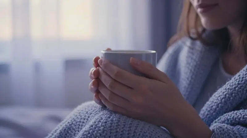 Manos sosteniendo una taza humeante en un dormitorio con luz suave, creando una atmósfera cálida y reconfortante.