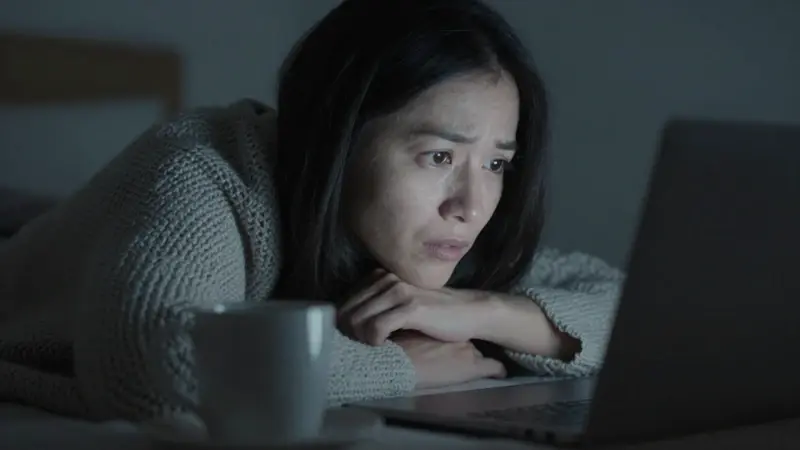 Mujer preocupada frente a un portátil en la oscuridad, posiblemente buscando respuestas sobre sus pesadillas. Ambiente nocturno y tenue con una taza de café cerca.